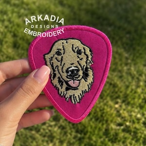 Puede incluir: Un parche bordado rosa en forma de púa de guitarra con la cara de un golden retriever. La cara del perro es marrón y negra, con una lengua rosa. El parche tiene el texto "ARKADIA DESIGNS EMBROIDERY" en blanco.