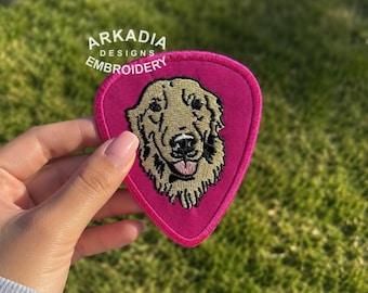 May include: A pink, guitar pick-shaped embroidered patch featuring a golden retriever's face. The dog's face is tan and black, with a pink tongue. The patch has the text "ARKADIA DESIGNS EMBROIDERY" in white.
