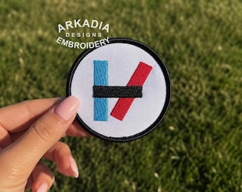 May include: Embroidered patch with a white background, featuring a blue vertical line, a red diagonal line, and a black horizontal line. The patch has a black border and the text "ARKADIA DESIGNS EMBROIDERY" in a circular design.