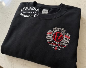 May include: A black crewneck sweatshirt featuring an embroidered design on the chest. The embroidery depicts the Ohio Stadium with a red Twenty One Pilots logo, the text "OHIO STADIUM", "OCT 17", "TWENTY ONE PILOTS", and an "O" emblem.