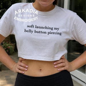 May include: A white short-sleeved crop top featuring the black embroidered text 'soft launching my belly button piercing' on the chest and a small blue, red, and white logo patch on the left sleeve. The shirt has a crew neckline and a cropped hem.