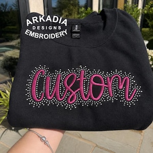 May include: A black crewneck sweatshirt featuring the word Custom embroidered in a dark pink script font. The text is surrounded by a dense cluster of small, shimmering silver rhinestones. The logo Arkadia Designs Embroidery is printed in white.