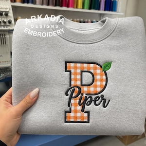 May include: A light grey crewneck sweatshirt featuring an embroidered design on the front. The design includes a large block letter P with an orange and white gingham pattern, a small green leaf, and the name Piper in black cursive script.