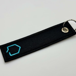 Puede incluir: Llavero de tela negro con una forma geométrica turquesa bordada. El llavero tiene un anillo de metal plateado.