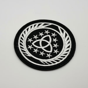 May include: A black and white embroidered patch with a circle design. The center of the circle features a white triquetra symbol surrounded by a ring of white stars. The outer ring of the circle is a white wreath design.