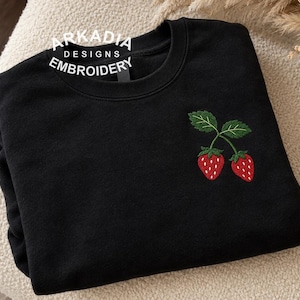 May include: A black crewneck sweatshirt featuring an embroidered design of two red strawberries with green leaves on the right chest. The text Arkadia Designs Embroidery is printed in white in a circular arc near the neckline.