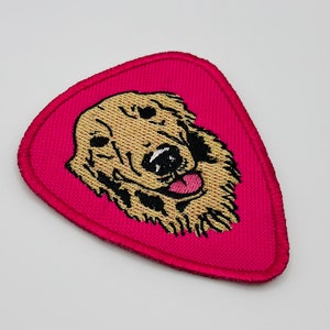 Op de afbeelding: Een roze gitaarplectrumvormige patch met een geborduurde afbeelding van een golden retriever hondenkop. De hond heeft zwarte ogen en een roze tong die uitsteekt.