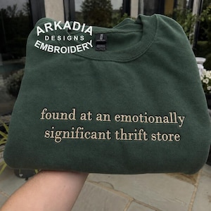 May include: A forest green crewneck sweatshirt featuring the embroidered text "found at an emotionally significant thrift store" in a light beige thread. The upper portion of the garment displays a white logo reading "ARKADIA DESIGNS EMBROIDERY".