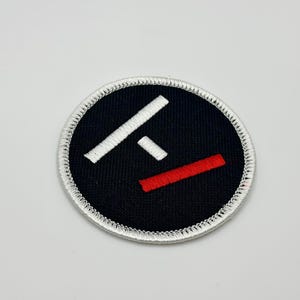 Op de afbeelding: Een geborduurde zwarte en witte patch met een witte "F" en een rode lijn eroverheen. De patch is rond en heeft een witte rand.