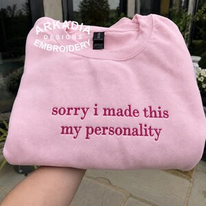 Op de afbeelding: Een lichtroze sweater met ronde hals en de geborduurde tekst "sorry i made this my personality" in donkerroze garen. Het kledingstuk is voorzien van een zwart neklabel en een wit "ARKADIA DESIGNS EMBROIDERY" watermerk in de hoek.