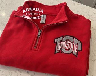 May include: A bright red quarter-zip pullover sweatshirt featuring a silver and white embroidered Ohio State logo on the chest. The garment includes a metal zipper closure and a white tag with the text Arkadia Designs Embroidery.