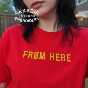 May include: A red t-shirt with the yellow text "FROM HERE". The shirt is a solid red colour and has short sleeves. The brand name "ARKADIA DESIGNS EMBROIDERY" is printed in white above the shirt.