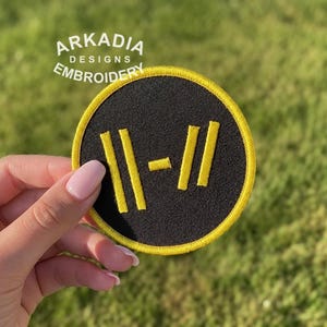 Puede incluir: Un parche bordado redondo negro con un borde amarillo y un diseño gráfico amarillo. El parche está sostenido por una mano sobre un fondo verde borroso. El texto "ARKADIA DESIGNS EMBROIDERY" es visible.