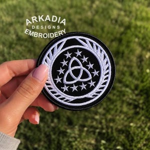 May include: A round, black embroidered patch with a white Celtic knot design surrounded by stars and a laurel wreath. The patch has the text "ARKADIA DESIGNS EMBROIDERY" in white.