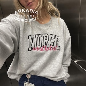May include: A grey crew neck sweatshirt with the word "NURSE" in black block letters and "anesthetist" in pink script. The sweatshirt also features the text "ARKADIA DESIGNS EMBROIDERY" in white. The sweatshirt is a casual style.
