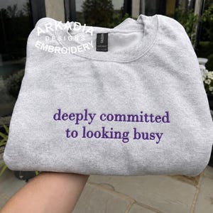 Könnte beinhalten: Ein hellgraues Sweatshirt mit Rundhalsausschnitt und dem violett gestickten Text "deeply committed to looking busy" mittig auf der Brust. Das Kleidungsstück hat ein schwarzes Nackenetikett mit dem Markennamen Gildan und ein weißes Logo-Wasserzeichen.