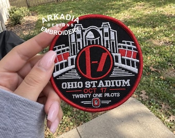Könnte beinhalten: Ein kreisförmiger gestickter Aufnäher mit schwarzem Hintergrund und rotem Rand. Das Design zeigt das Ohio Stadium in weißem und rotem Garn, einen zentralen roten Kreis mit dem schwarzen Twenty One Pilots Logo sowie den Text OHIO STADIUM, OCT 17, TWENTY ONE PILOTS.