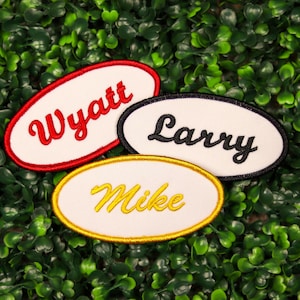May include: Three embroidered name patches with white ovals and colorful borders. The names "Wyatt" in red, "Larry" in black, and "Mike" in yellow are stitched on the patches. The patches are set against a green background.