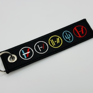 Peut inclure: Porte-clés brodé noir avec cinq motifs circulaires colorés. Chaque motif présente un symbole unique en rouge, bleu, jaune et turquoise. Le porte-clés comprend un anneau en métal argenté pour attacher aux clés ou aux sacs.