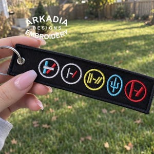 May include: A black rectangular embroidered keychain featuring five circular symbols in a row. The symbols are stitched in white, yellow, light blue, and red thread. A silver metal split ring is attached to the left side of the fabric tag.