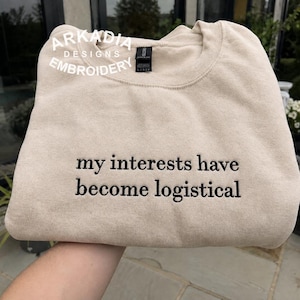 Puede incluir: Una sudadera beige de cuello redondo con texto bordado en negro que dice "my interests have become logistical". La prenda incluye un logotipo blanco en la esquina superior izquierda que dice "ARKADIA DESIGNS EMBROIDERY".