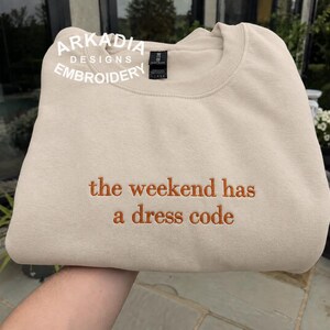 Może przedstawiać: Beżowa bluza z okrągłym dekoltem i wyhaftowanym napisem "the weekend has a dress code" wykonanym nicią w kolorze rdzy. Odzież posiada czarną metkę na karku oraz biały znak wodny w lewym górnym rogu z napisem "ARKADIA DESIGNS EMBROIDERY".