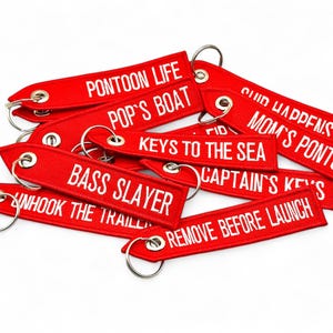 May include: A selection of red fabric keyrings with white embroidered text. Phrases include "Pontoon Life," "Bass Slayer," and "Remove Before Launch." Each keyring has a metal ring for attachment.