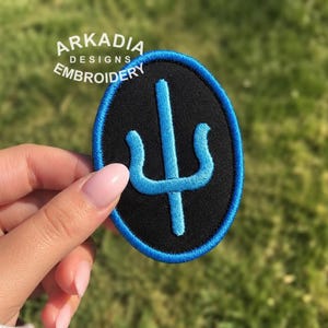 Puede incluir: Un parche bordado con una base ovalada negra y un borde azul. Un símbolo azul está centrado en el parche. El texto "ARKADIA DESIGNS EMBROIDERY" está arqueado sobre el parche.