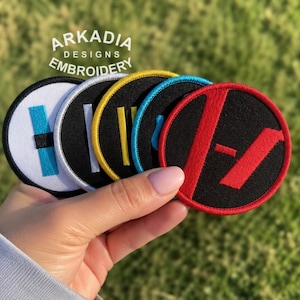 以下が含まれることがあります： さまざまなデザインの丸い刺繍パッチが5つ。パッチは、黒、白、黄色、赤の背景にコントラストのあるシンボルが特徴です。背景はぼやけた緑色です。「ARKADIA DESIGNS EMBROIDERY」の文字が見えます。