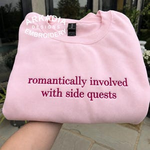 Könnte beinhalten: Ein hellrosa Sweatshirt mit Rundhalsausschnitt und dem gestickten Text "romantically involved with side quests" in dunkelrosa Garn. Das Kleidungsstück hat ein schwarzes Nackenetikett und ein weißes Logo mit "ARKADIA DESIGNS EMBROIDERY".