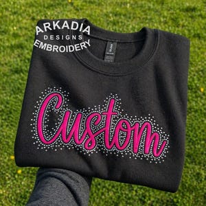 May include: Black sweatshirt with the word "Custom" in pink script, outlined with small white dots. The text "ARKADIA DESIGNS EMBROIDERY" is above the word "Custom". The sweatshirt is displayed on a green grassy background.