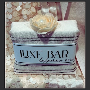 May include: A white bar of soap with a black and white striped design. The soap is decorated with a white flower and has a label that reads "LUXE BAR bulgarian rose".