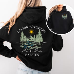 May include: Black hoodie with a landscape design on the back, including trees, a river, mountains, and a sun. The text reads "LIFETIME ADVENTURE" above the design, with "Mr. & Mrs. BARSTEN" below. A smaller version of the design is on the front.