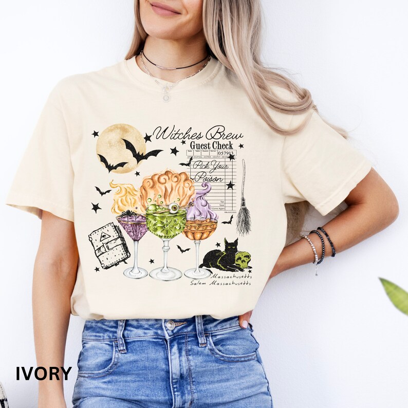 May include: A beige t-shirt with a graphic design of a witch's brew with a black cat, a broom, and a guest check. The text on the shirt reads "Witches Brew Guest Check Pick Your Poison Salem Massachusetts".