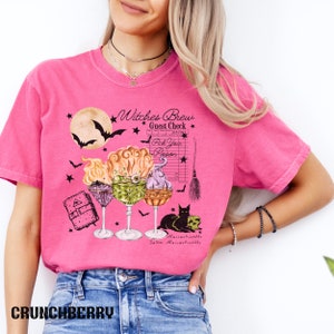 May include: Pink t-shirt with a graphic of three witches' brew cocktails, a black cat, a skull, a broom, a book, and a moon with bats. The text on the shirt reads "Witches Brew Guest Check Pick Your Poison Salem Massachusetts".