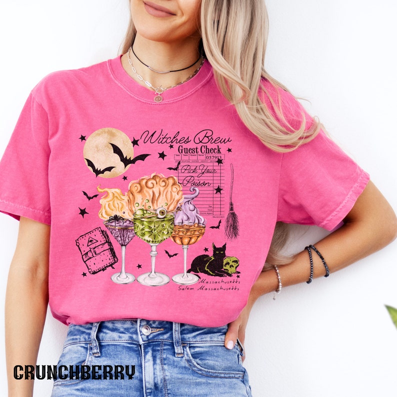 May include: Pink t-shirt with a graphic of three witches' brew cocktails, a black cat, a skull, a broom, a book, and a moon with bats. The text on the shirt reads "Witches Brew Guest Check Pick Your Poison Salem Massachusetts".