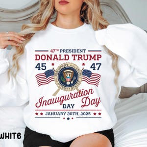 以下が含まれることがあります： 白のトレーナーに、大統領の紋章と2つのアメリカ国旗を配したグラフィックデザインが施されています。デザインには「47th President Donald Trump 45 47」というテキストと、「Inauguration Day January 20th, 2025」というテキストも含まれています。