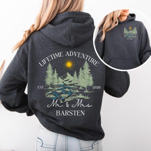 May include: Charcoal grey hooded sweatshirt with a front pocket. The back displays a graphic with the words "LIFETIME ADVENTURE" above a mountain and river scene, with "EST. 2026" and "Mr. & Mrs. BARSTEN" below. A smaller version of the graphic is on the front.