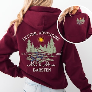 May include: Burgundy hoodie with a landscape design featuring trees, mountains, a river, and a sun. The back of the hoodie reads "LIFETIME ADVENTURE" above the landscape, with "Mr. & Mrs. BARSTEN" below. The front has a smaller version of the design.