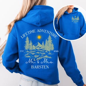 May include: A royal blue hoodie featuring a nature-themed design. The graphic includes a sun, mountains, trees, and the text "LIFETIME ADVENTURE" and "Mr. & Mrs. BARSTEN". The year "2026" is also displayed.