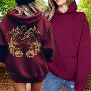May include: Two maroon hoodies, one with a gold mountain scene graphic on the back. The graphic features mountains, trees, a lake, and a sun.