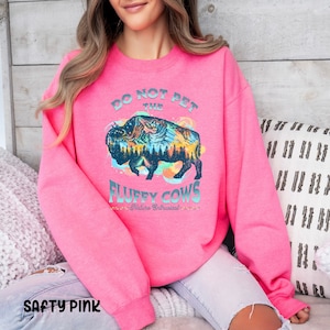Pu&ograve; includere: Una felpa rosa con il testo "Do Not Pet The Fluffy Cows - Nature Enthusiast" e una grafica di un bisonte con una scena di montagna al suo interno.