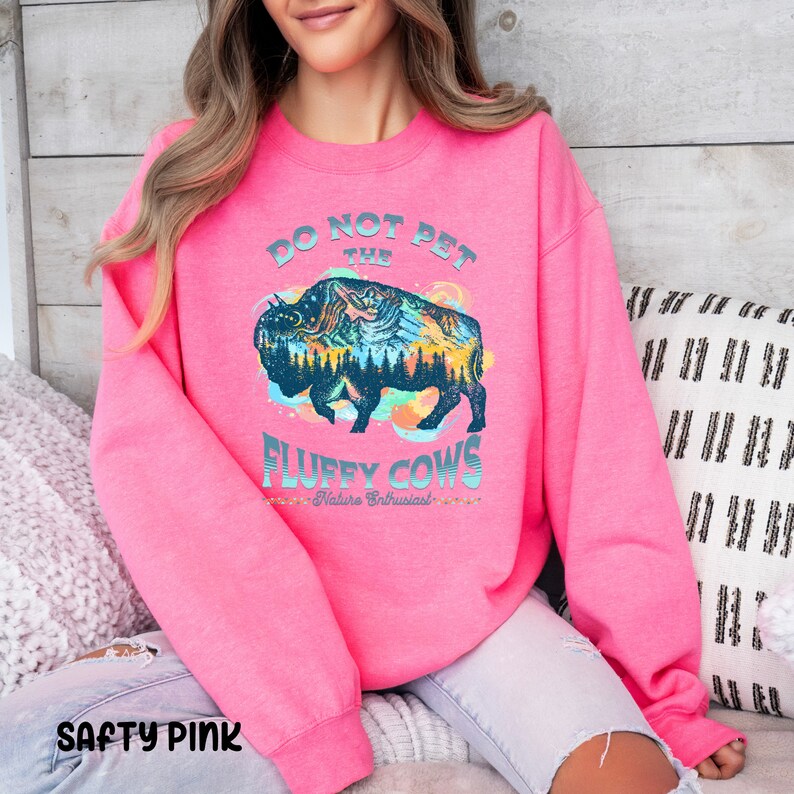 Pu&ograve; includere: Una felpa rosa con il testo "Do Not Pet The Fluffy Cows - Nature Enthusiast" e una grafica di un bisonte con una scena di montagna al suo interno.