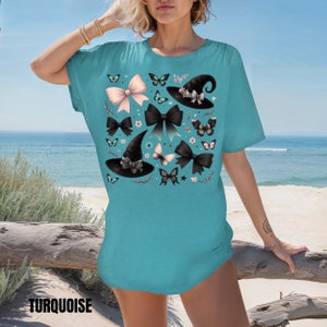 Pode incluir: Uma camiseta azul turquesa com uma estampa de chap&eacute;u de bruxa, la&ccedil;o e borboleta. A estampa apresenta chap&eacute;us de bruxa pretos, la&ccedil;os rosa e pretos e borboletas pretas.