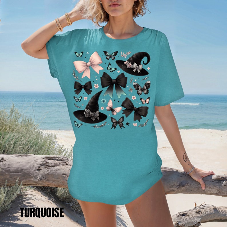 Pode incluir: Uma camiseta azul turquesa com uma estampa de chap&eacute;u de bruxa, la&ccedil;o e borboleta. A estampa apresenta chap&eacute;us de bruxa pretos, la&ccedil;os rosa e pretos e borboletas pretas.