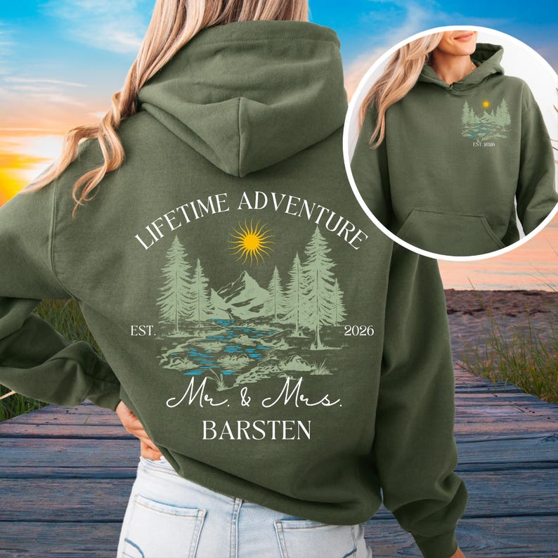 May include: An olive green hoodie with a landscape design featuring trees, mountains, a river, and a sun. The text "LIFETIME ADVENTURE" arches above the design, with "Mr. & Mrs. BARSTEN" below. The year "2026" is also printed on the hoodie.