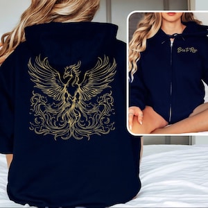 Könnte beinhalten: Marineblauer Kapuzenpullover mit Reißverschluss und goldenem Phönix-Design auf dem Rücken. Der Phönix hat ausgebreitete Flügel und detaillierte Merkmale. Auf der Vorderseite des Hoodies steht in Gold der Text "Born to Rise".