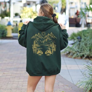 May include: A dark green hooded sweatshirt with a gold mountain scene on the back. The scene includes mountains, a lake, trees, and a sun.