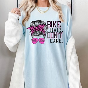 Puede incluir: Camiseta azul claro con un gr&aacute;fico de una mujer con un mo&ntilde;o desordenado, una diadema de leopardo rosa y gafas de sol. El texto "Bike Hair. Don't Care." est&aacute; impreso en la camiseta.