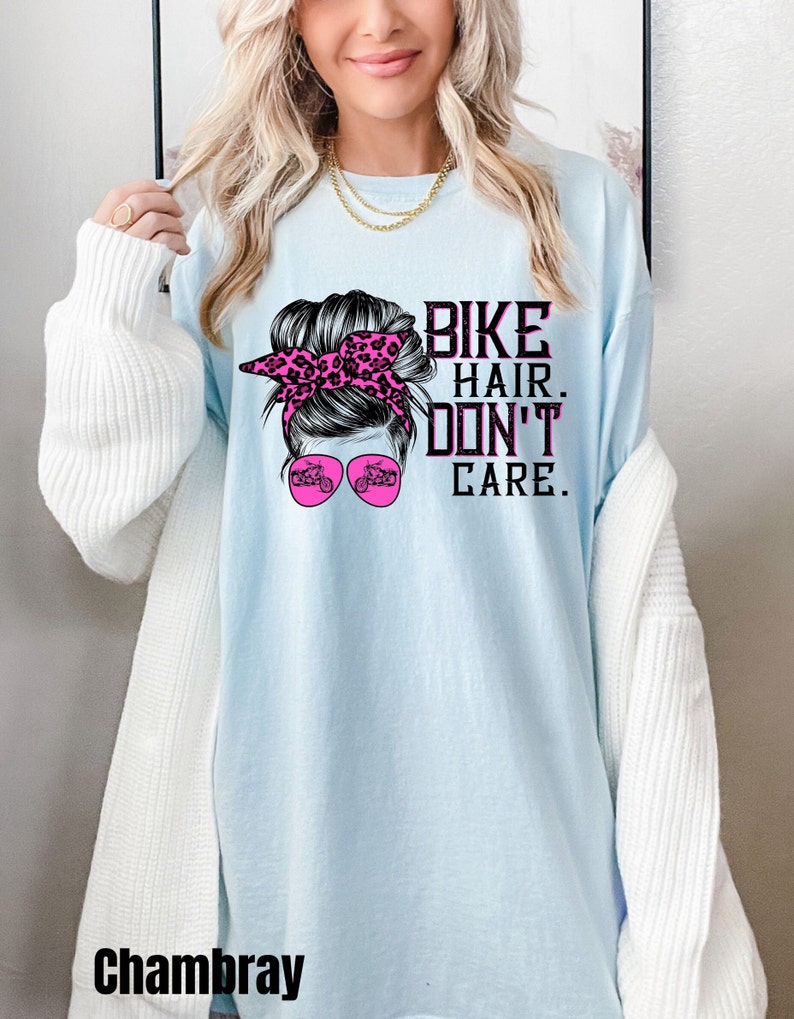 Puede incluir: Camiseta azul claro con un gr&aacute;fico de una mujer con un mo&ntilde;o desordenado, una diadema de leopardo rosa y gafas de sol. El texto "Bike Hair. Don't Care." est&aacute; impreso en la camiseta.